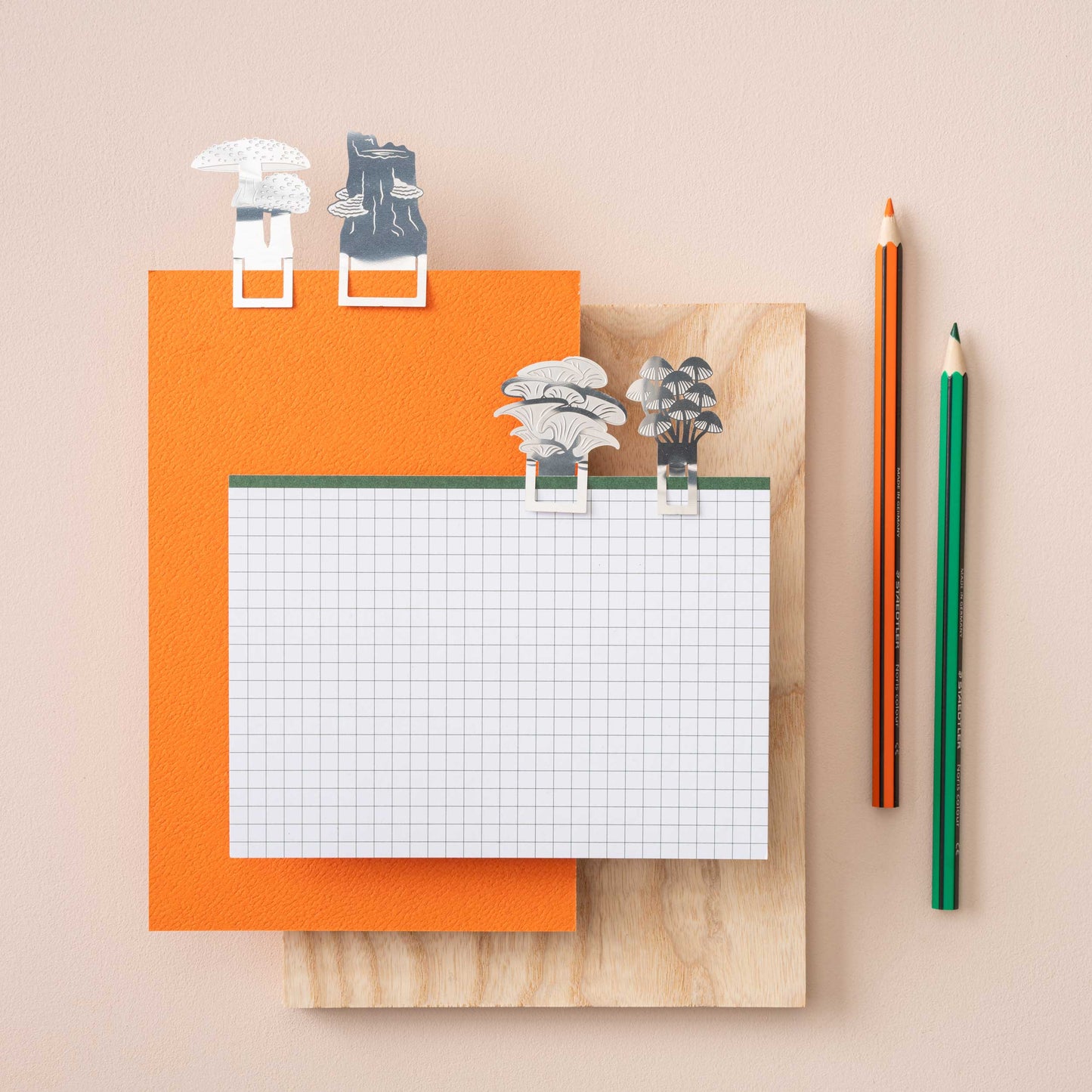 A set of stainless steel mushroom bookmarks in various designs, displayed on top of a notebook with a pen and decorative elements by Another Studio