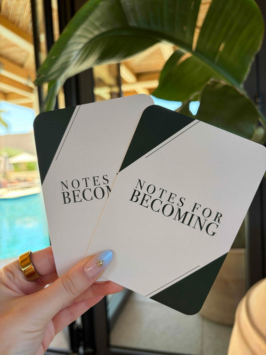 A hand holding two white and dark green cards printed with the text 'NOTES FOR BECOMING' in black serif font, with a blurred resort-style background featuring a pool and large green leaves.