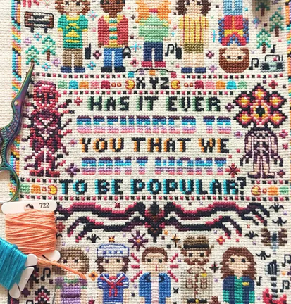 Cross-stitched pattern with text and character illustrations, accompanied by a spool of thread and scissors.
