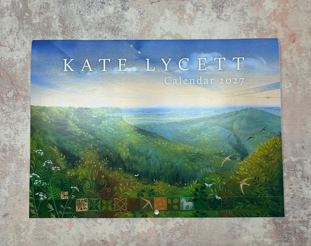 Kate Lycett 2027 Calendar featuring a landscape painting of rolling green hills under a blue sky, displayed flat on a textured gray surface.