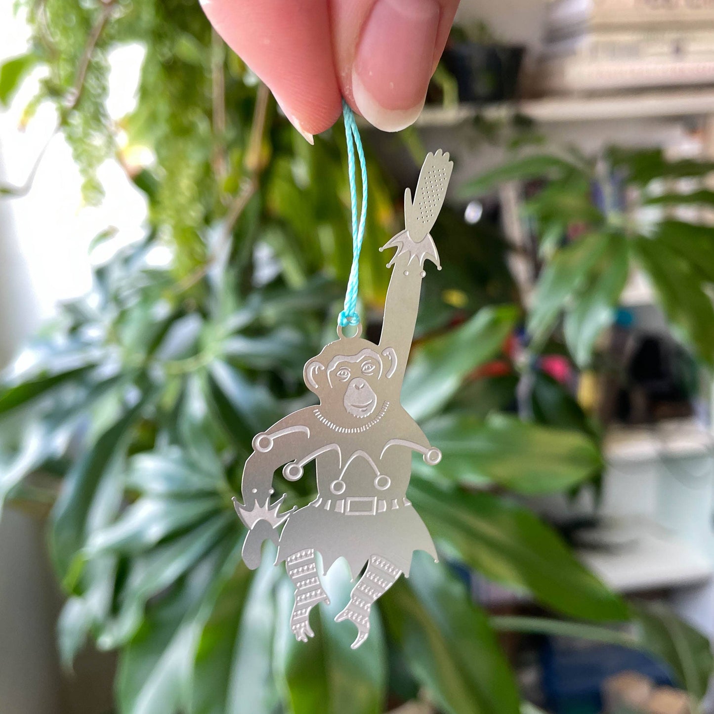 A stainless steel chimpanzee-shaped Christmas ornament hanging from a blue cord, displayed against a plant with green leaves in the background by Another Studio for Heart Gallery