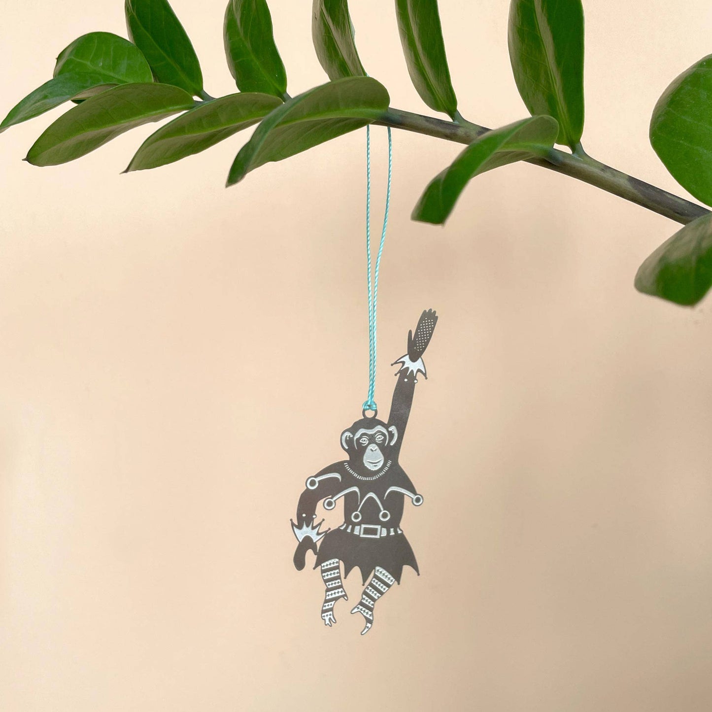 A stainless steel chimpanzee-shaped Christmas ornament hanging from a blue cord, displayed against a plant with green leaves in the background by Another Studio for Heart Gallery