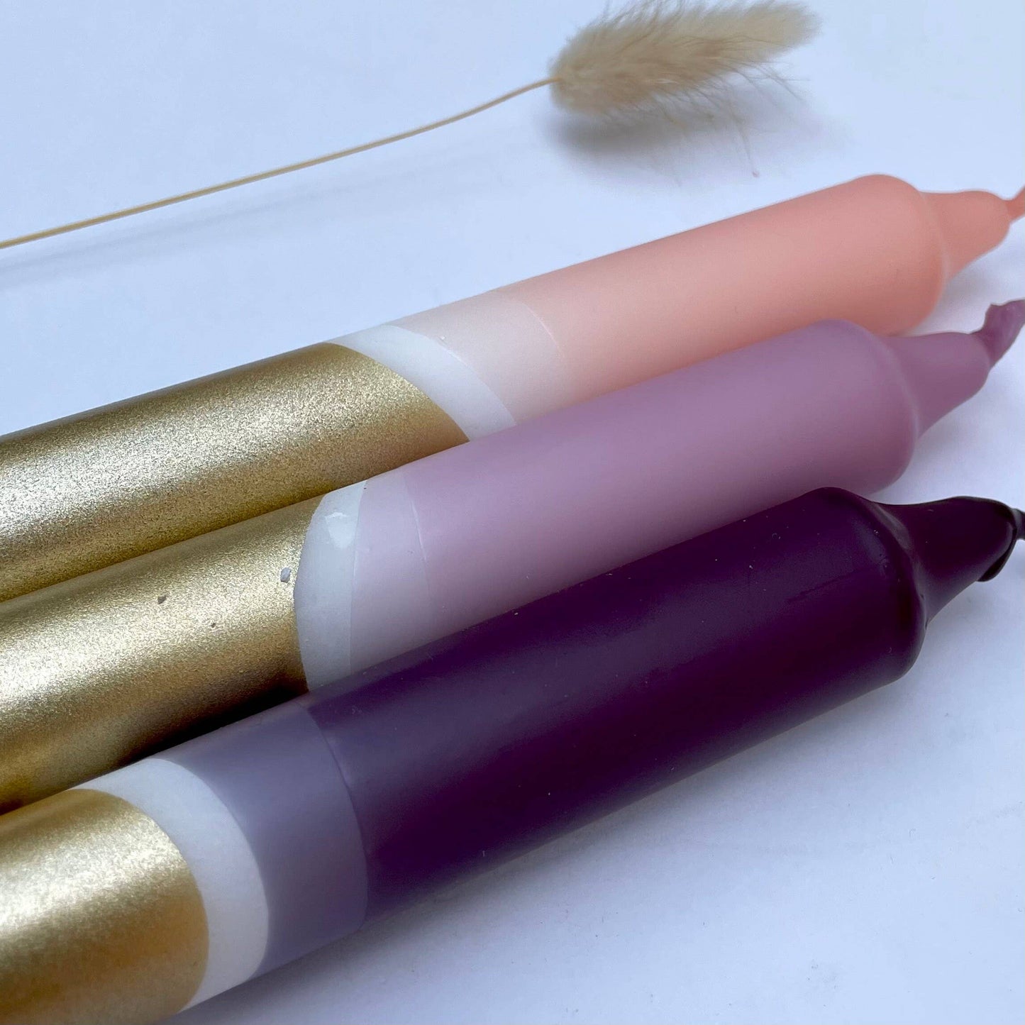 close up of a  set of three hand-dipped dinner candles in aubergine, heather, and blush pink with gold accents, displayed alongside a dried plant on a white background by The Singing Rabbit