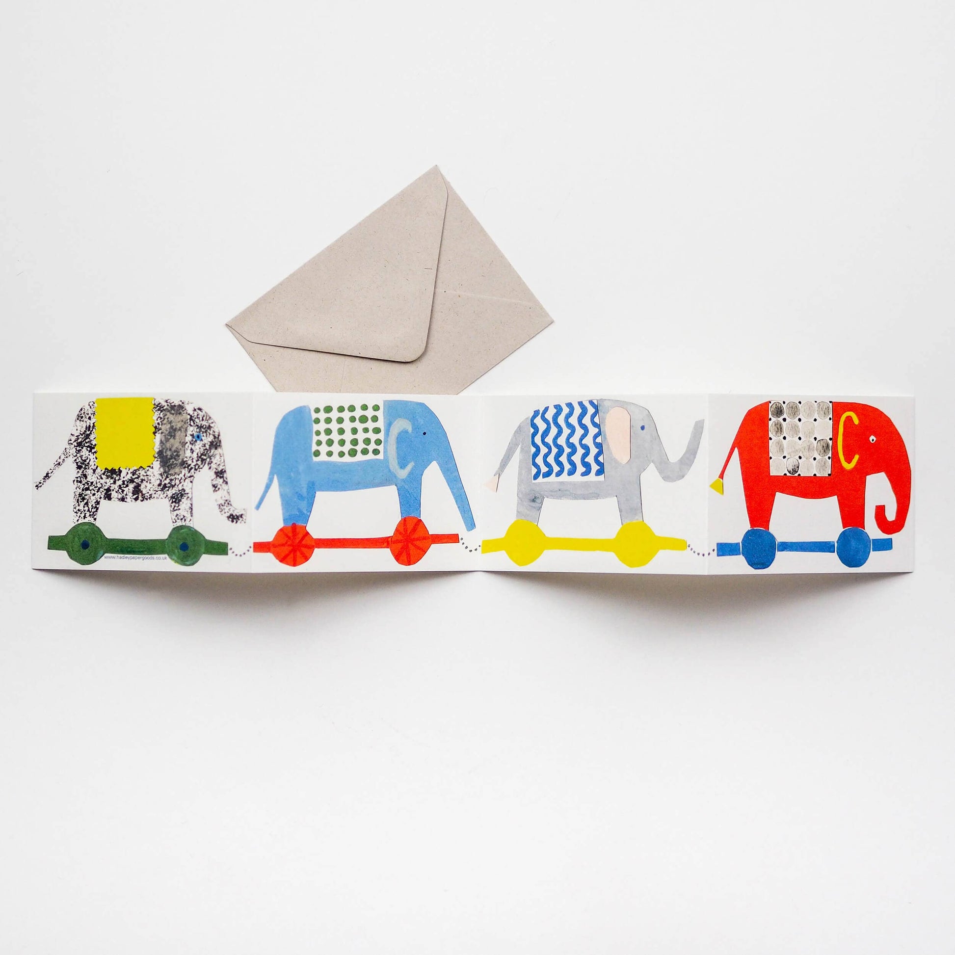 Colorful card  by Hadley with four elephants on wheels and a beige envelope on a white background