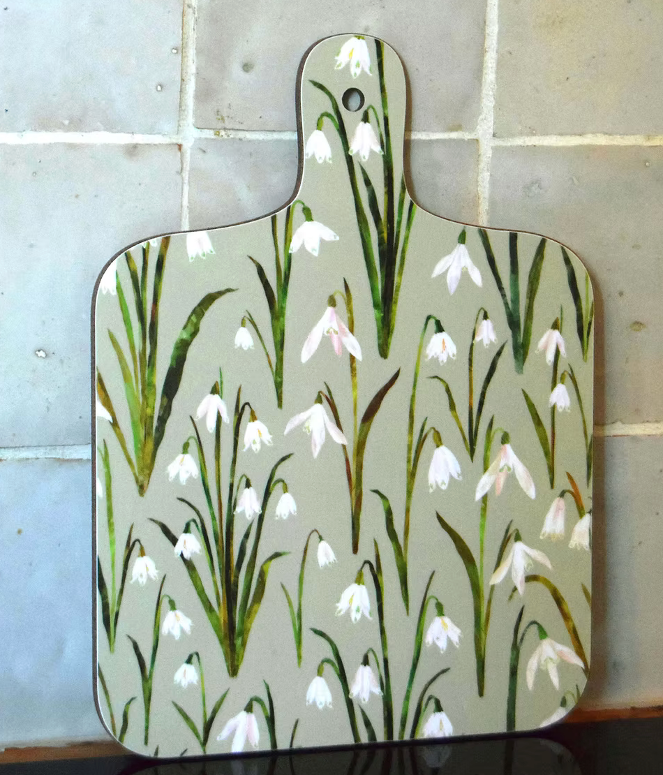 A decorative mini chopping board with a lovely snowdropdesign  on a white background, displayed on a wooden surface with vegetables around it.