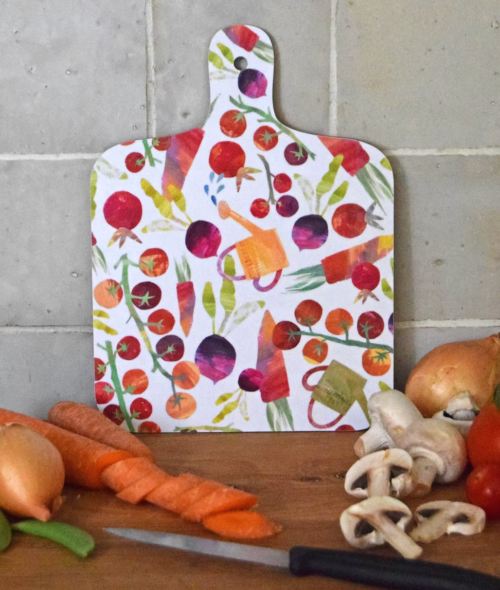 A decorative mini chopping board with a colorful busy veg pattern on a white background, displayed on a wooden surface with vegetables around it.