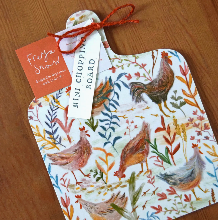 A decorative mini chopping board with a colorful chicken floral pattern on a white background, displayed on a wooden surface with vegetables around it.
