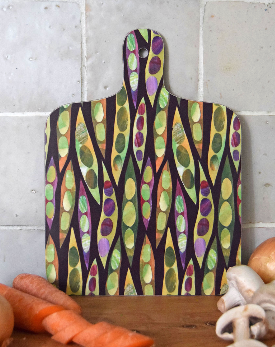 A decorative mini chopping board with a colorful pea pattern on a white background, displayed on a wooden surface with vegetables around it. Heart Gallery