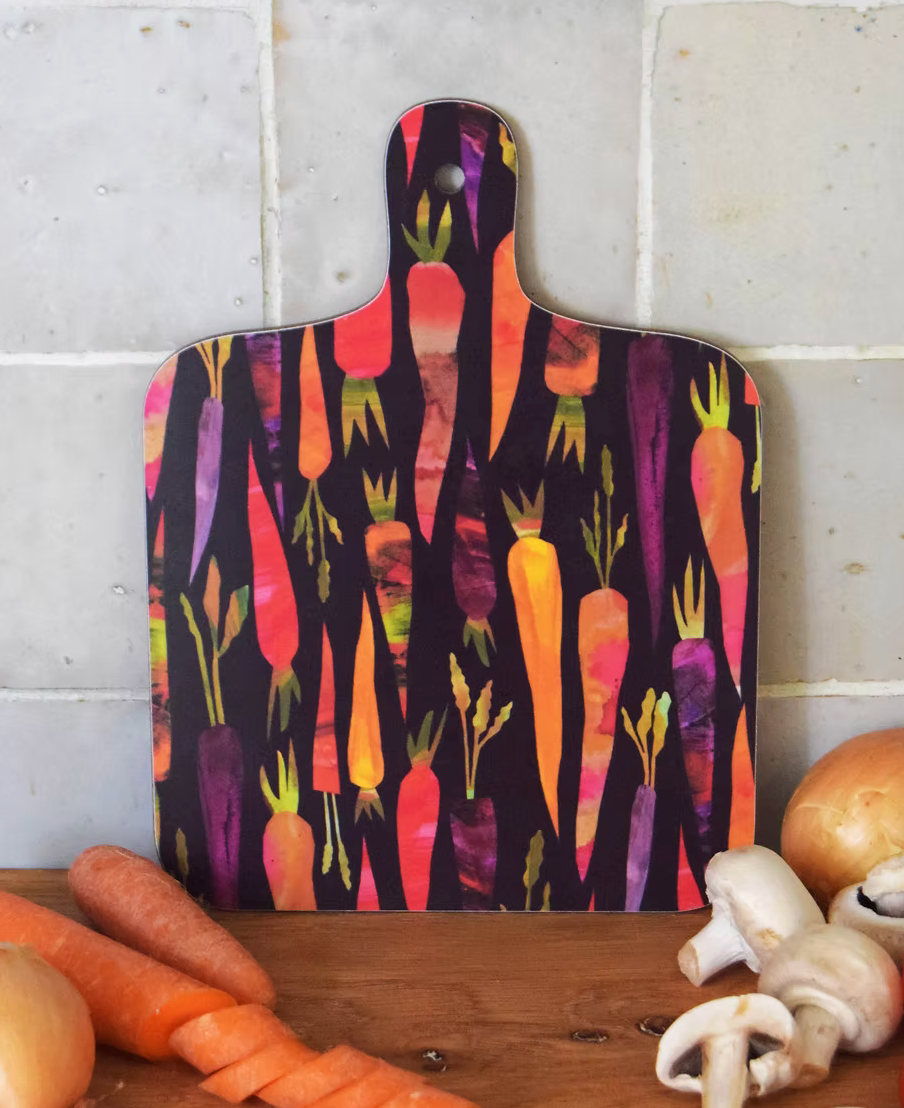 A decorative mini chopping board with a purple carrot pattern on a white background, displayed on a wooden surface with vegetables around it.