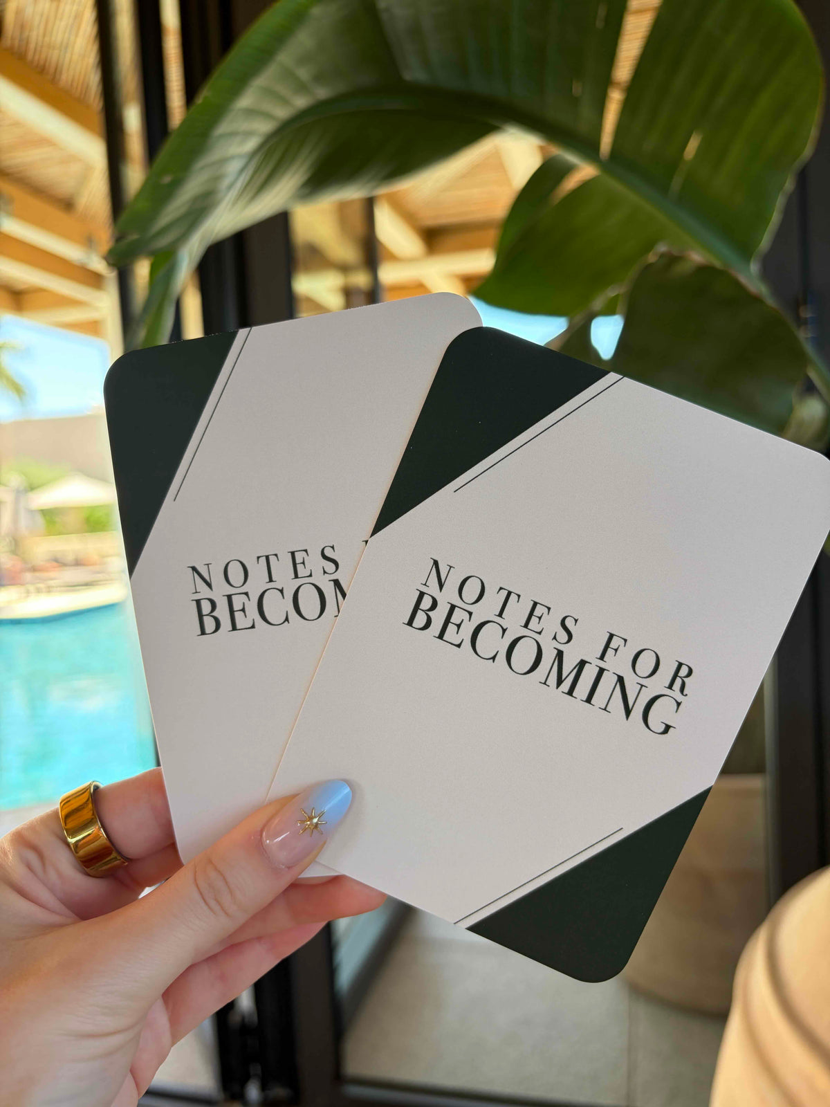A hand holding two white and dark green cards printed with the text 'NOTES FOR BECOMING' in black serif font, with a blurred resort-style background featuring a pool and large green leaves.