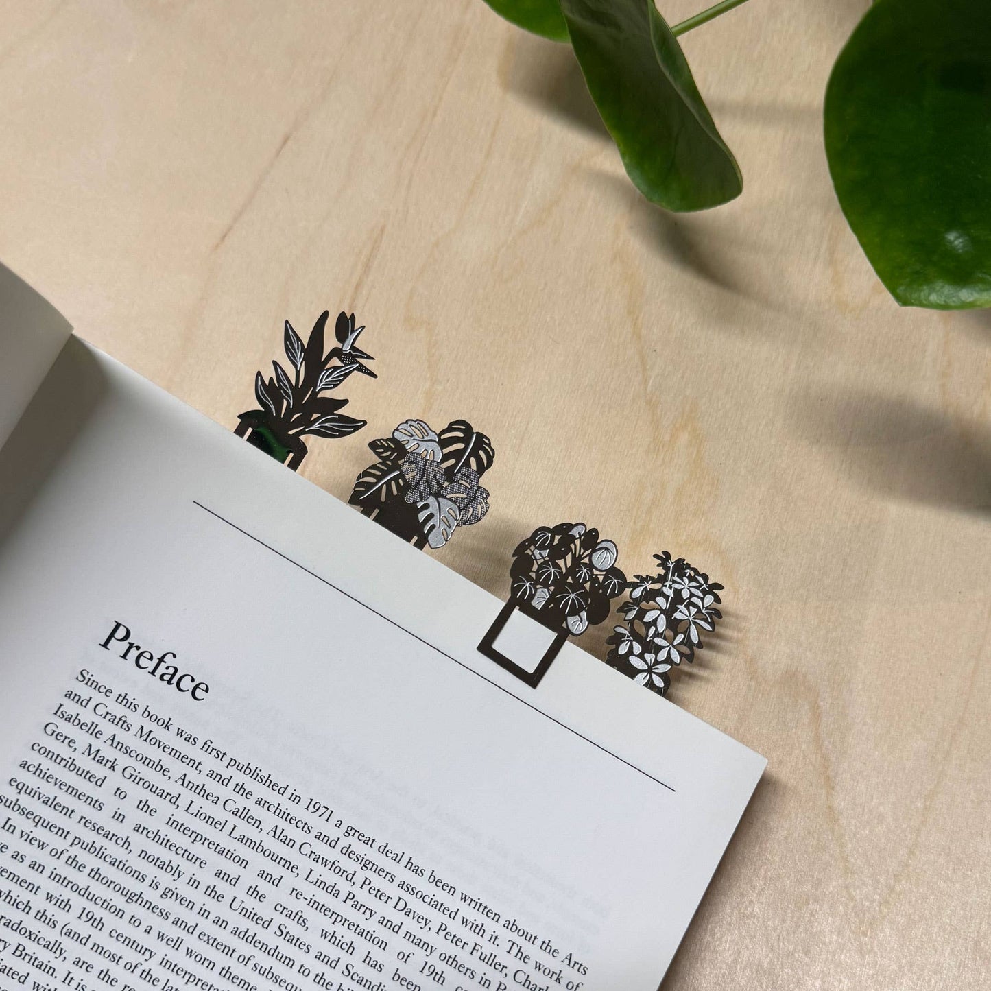 A set of four stainless steel bookmarks with a botanical design, featuring illustrations of houseplants such as Monstera delicious, Pilea peperomioides, Strelitzia, and Schefflera, presented on eco-friendly packaging by Another Studio for Heart Gallery