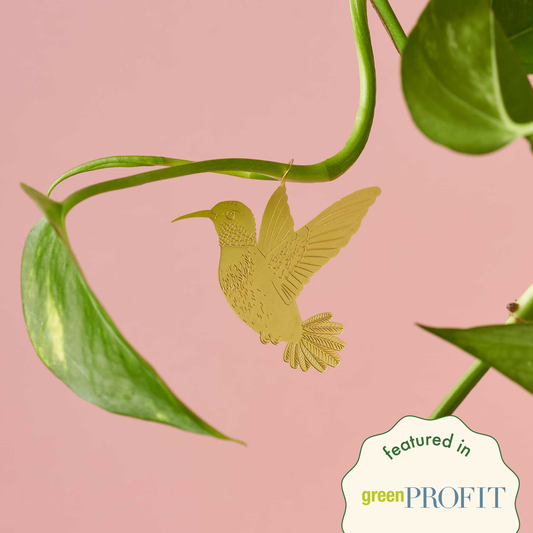 A gold-colored metal hummingbird decoration attached to a green plant leaf against a pink background by Another Studio for Heart Gallery