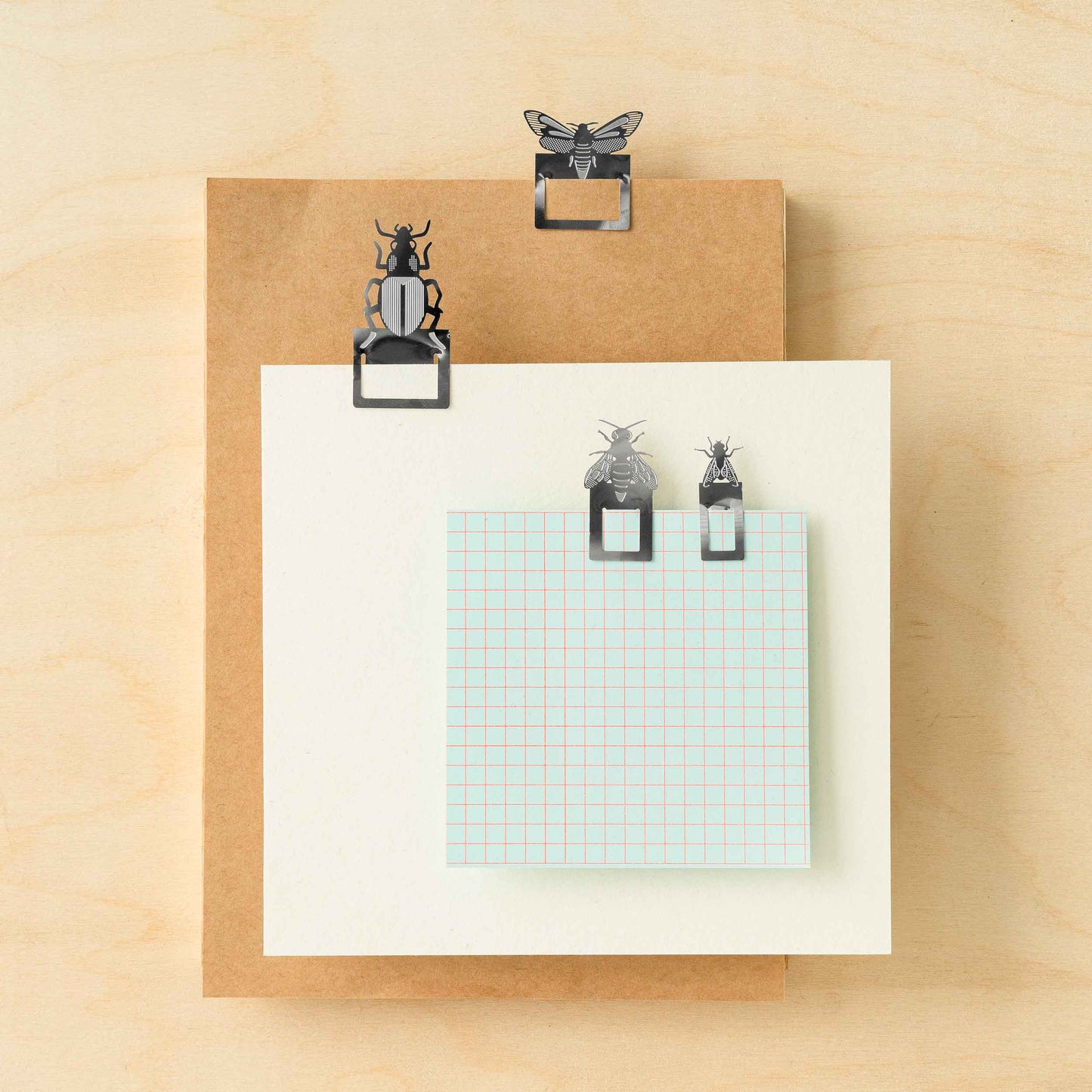 A set of metal page markers in the shapes of insects such as a moth and fly, attached to a paper clip, displayed on a wooden background with a piece of paper showing grid lines.