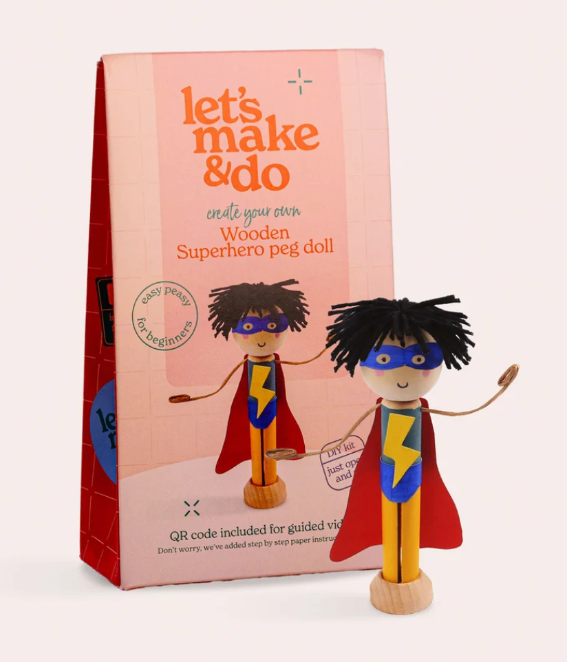 A wooden superhero peg doll with a black yarn hair, blue mask, yellow lightning-bolt chest, and red cape, standing next to its pink packaging box labeled 'let's make & do'.