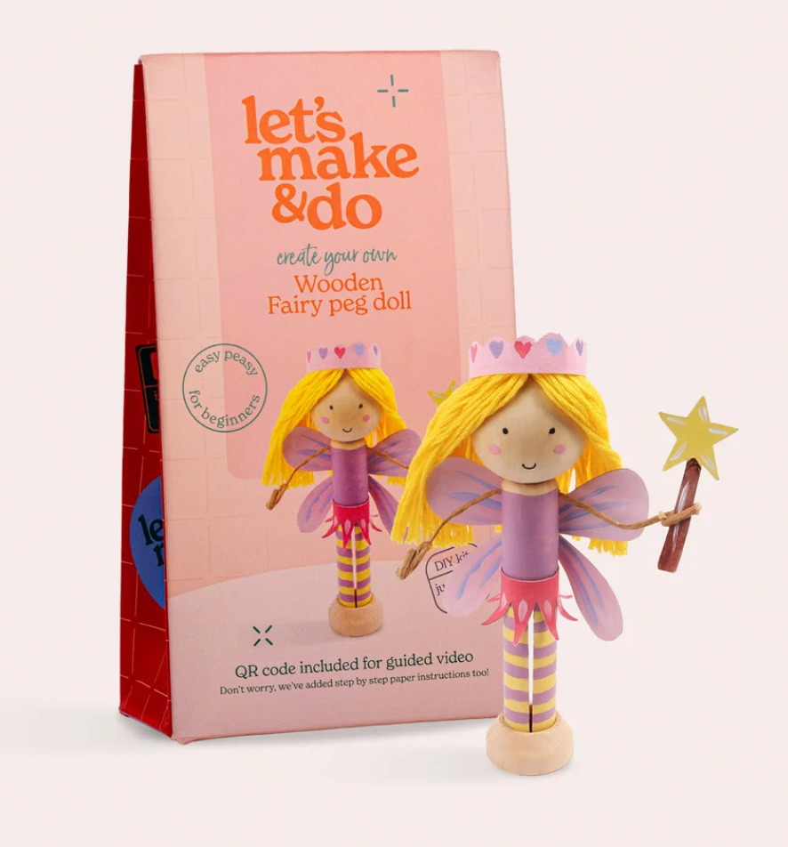 A wooden fairy peg doll with yellow hair, purple wings, and a star wand stands in front of its pink packaging box on a light beige background.