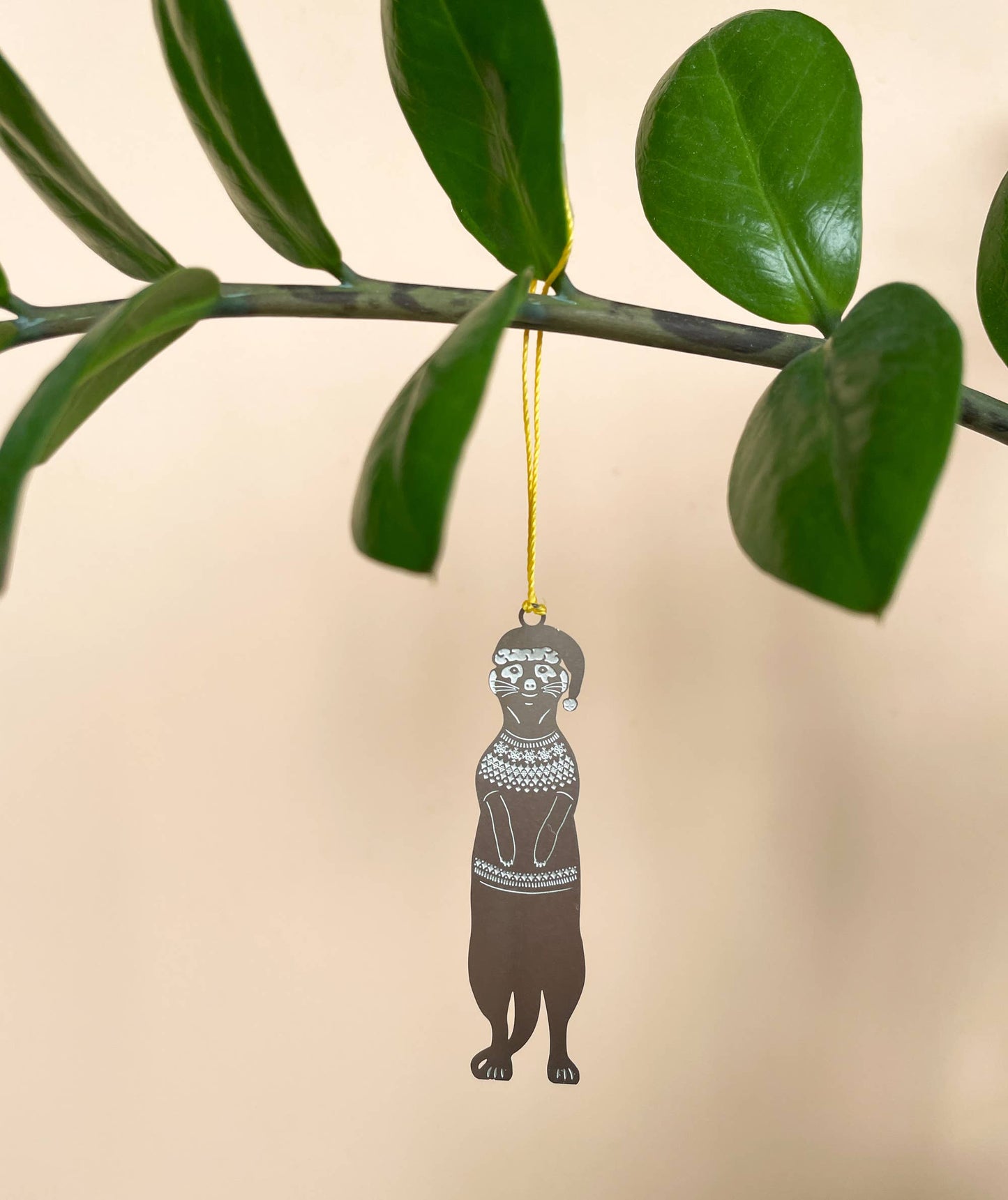 A stainless steel meerkat Christmas jumper decoration with a festive color cord, hanging on a green leaf.