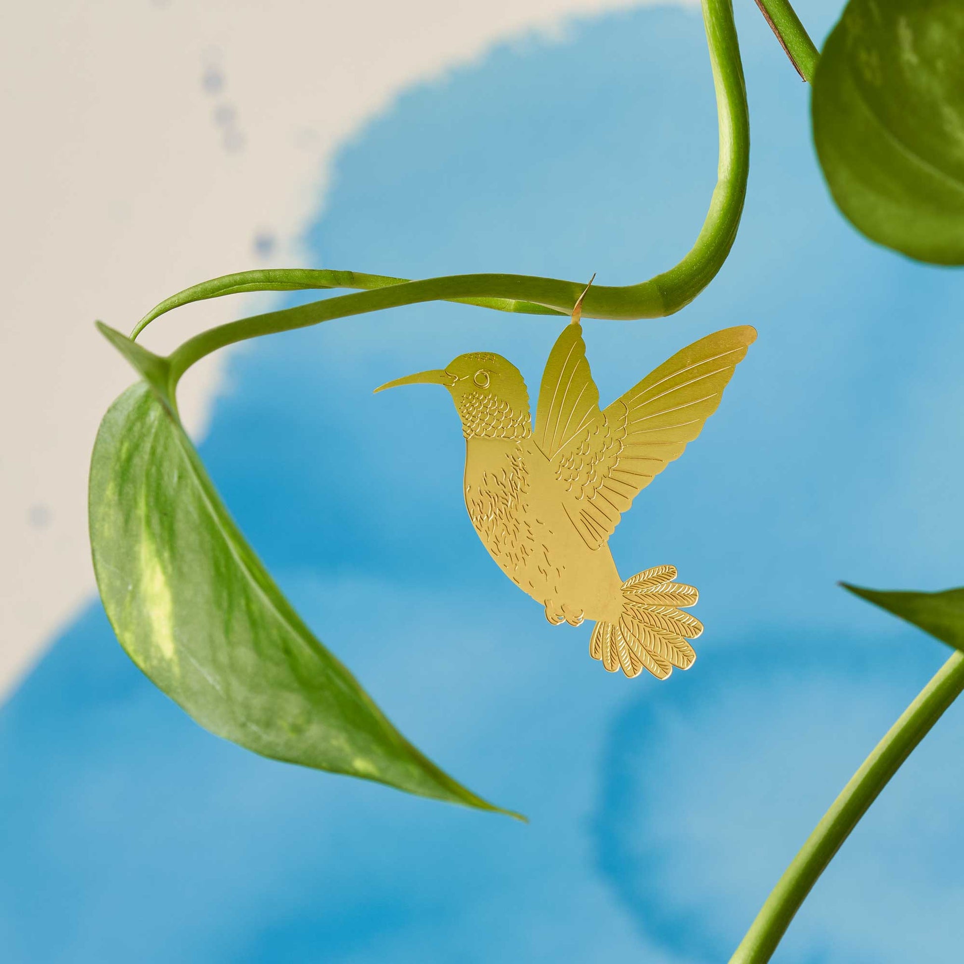 A gold-colored metal hummingbird decoration attached to a green plant leaf against a blue background by Another Studio for Heart Gallery