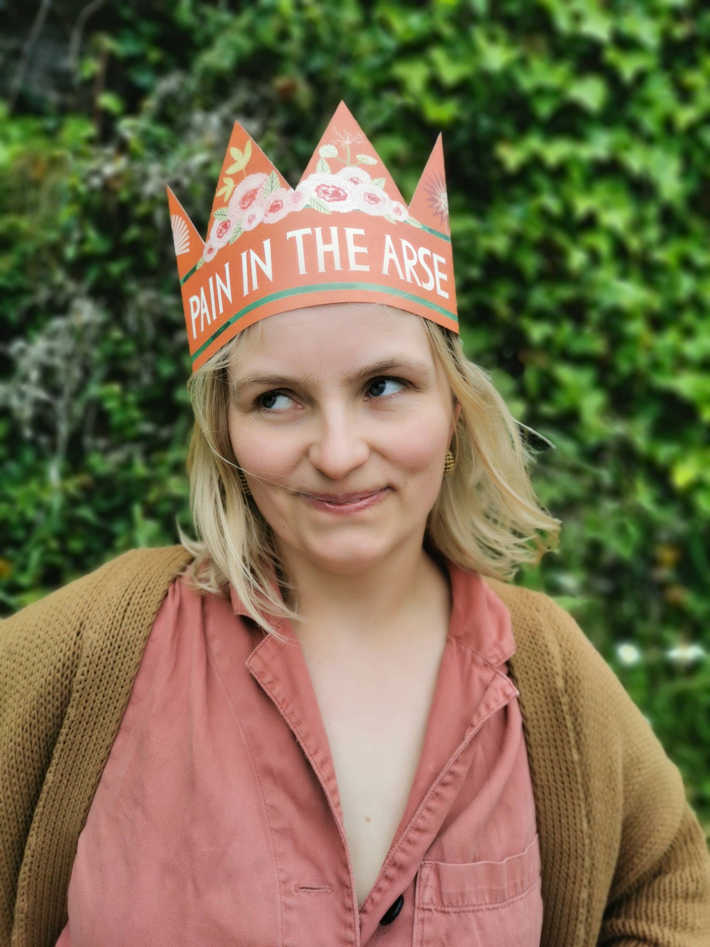 Person wearing a paper crown with text, standing against a green hedge. Pain in the ARse crown, birthday card