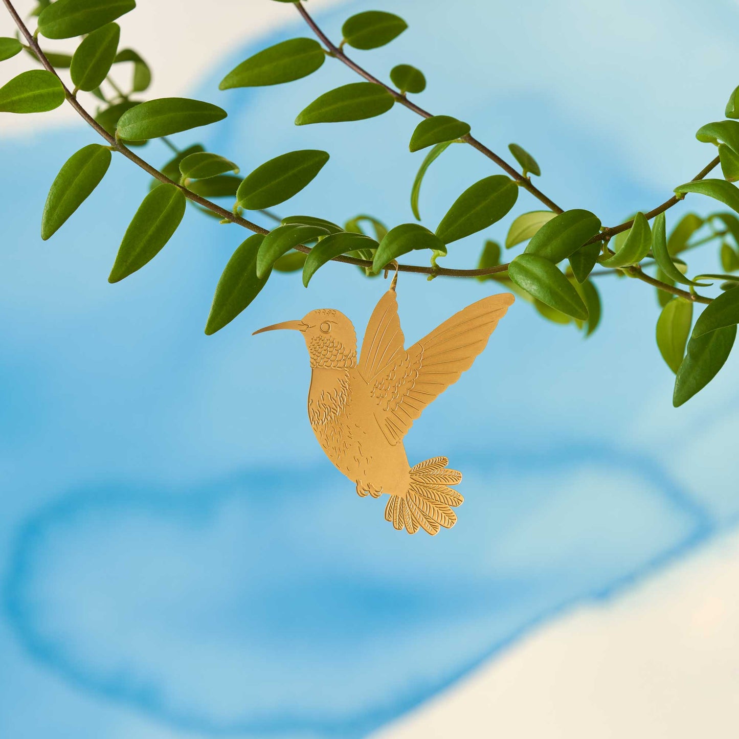 A gold-colored metal hummingbird decoration attached to a green plant leaf against a blue background by Another Studio for Heart Gallery