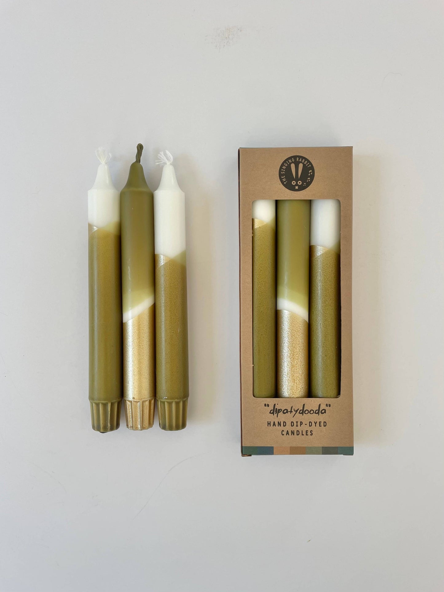 A set of three dip-dyed dinner candles with olive green and gold colors, packaged in a box with a transparent window displaying the candles inside.