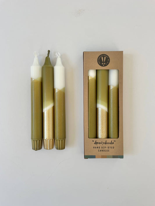 A set of three dip-dyed dinner candles with olive green and gold colors, packaged in a box with a transparent window displaying the candles inside.