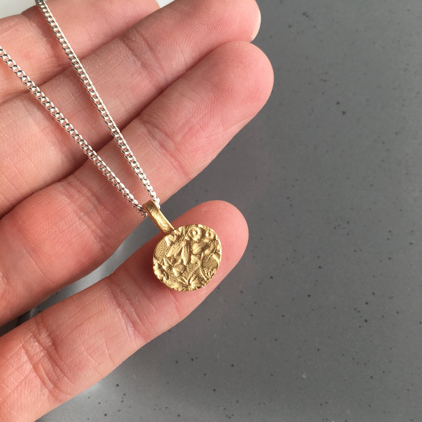 Gold pendant on a silver chain held between fingers against a gray background