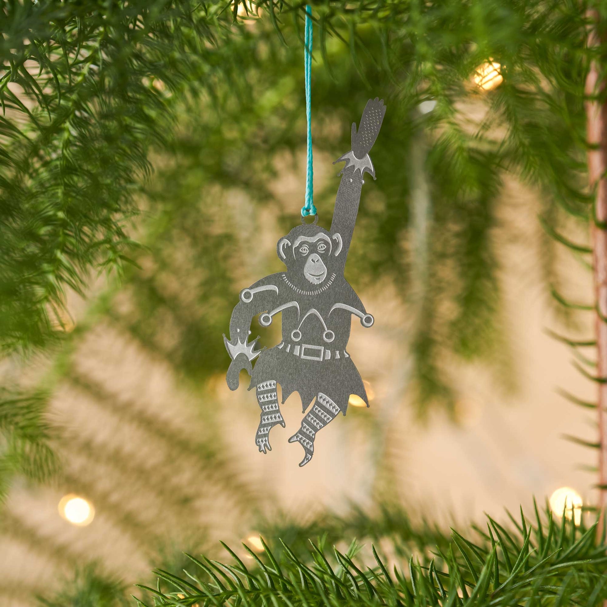 A stainless steel chimpanzee-shaped Christmas ornament hanging from a blue cord, displayed against a plant with a Christmas Tree  in the background by Another Studio for Heart Gallery