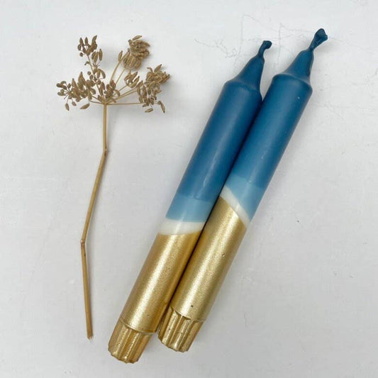 Two dinner candles with a dip dyed design  in teal blue and gold, alongside a dried flower decoration.