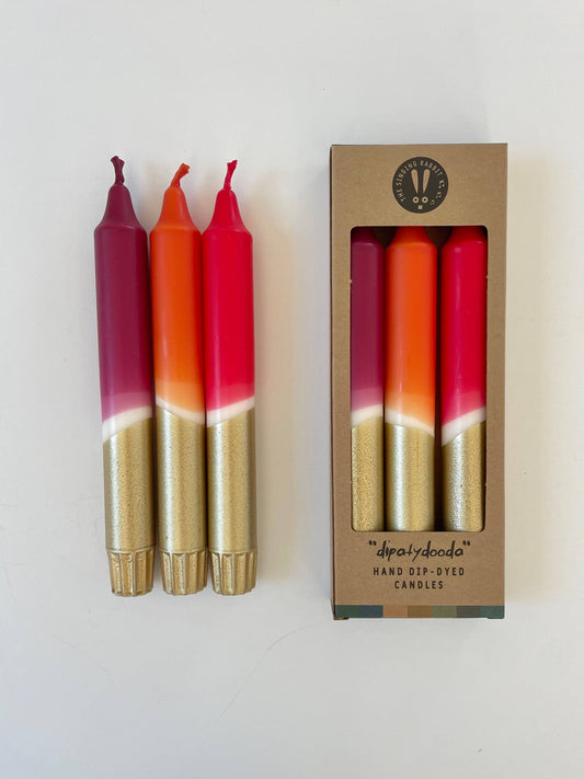 Three dip-dyed candles with gold and red gradient in front of their packaging box.