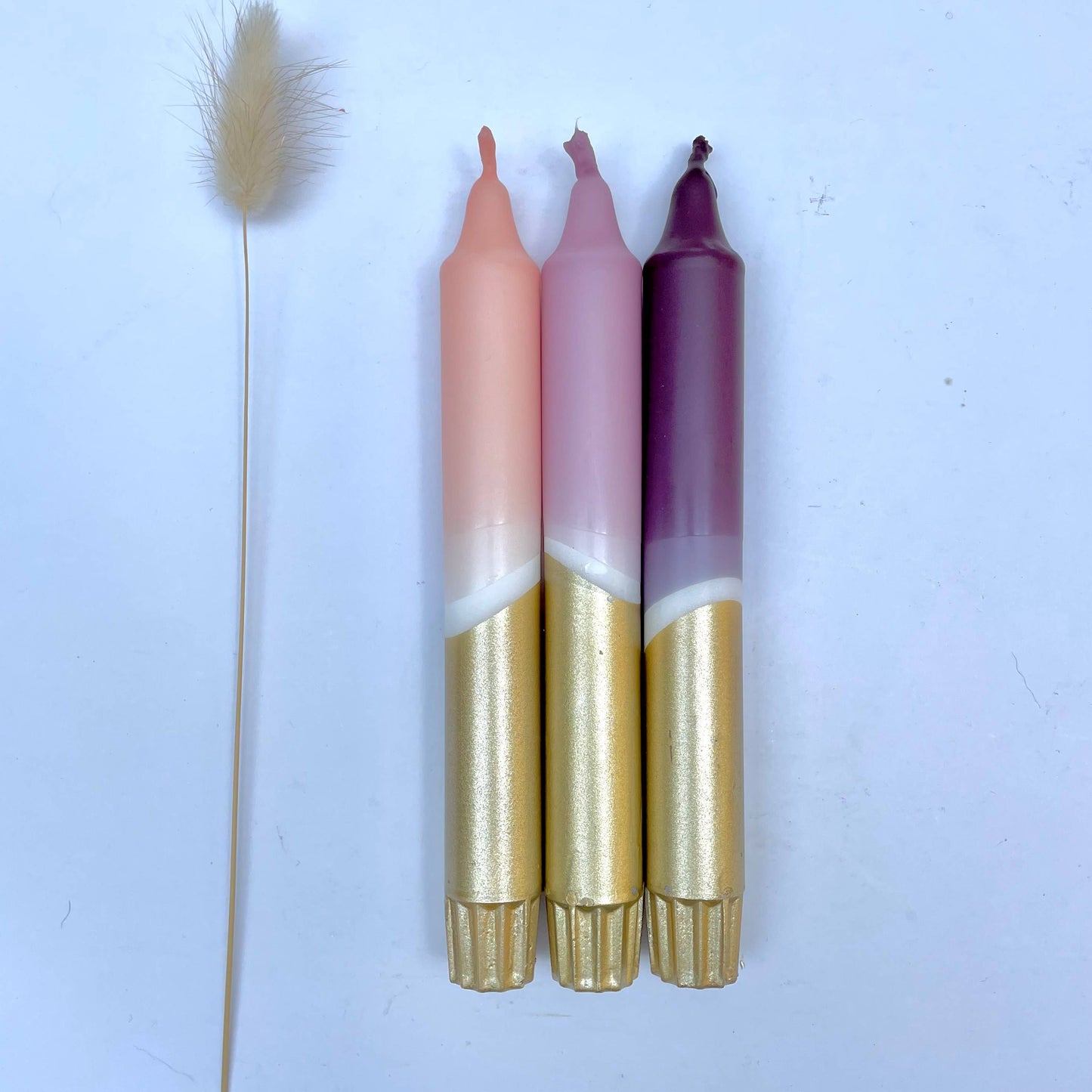 A set of three hand-dipped dinner candles in aubergine, heather, and blush pink with gold accents, displayed alongside a dried plant on a white background.