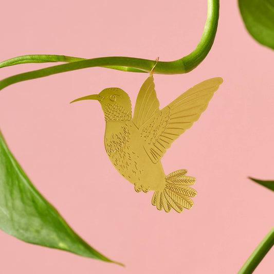 A gold-colored metal hummingbird decoration attached to a green plant leaf against a pink background by Another Studio for Heart Gallery