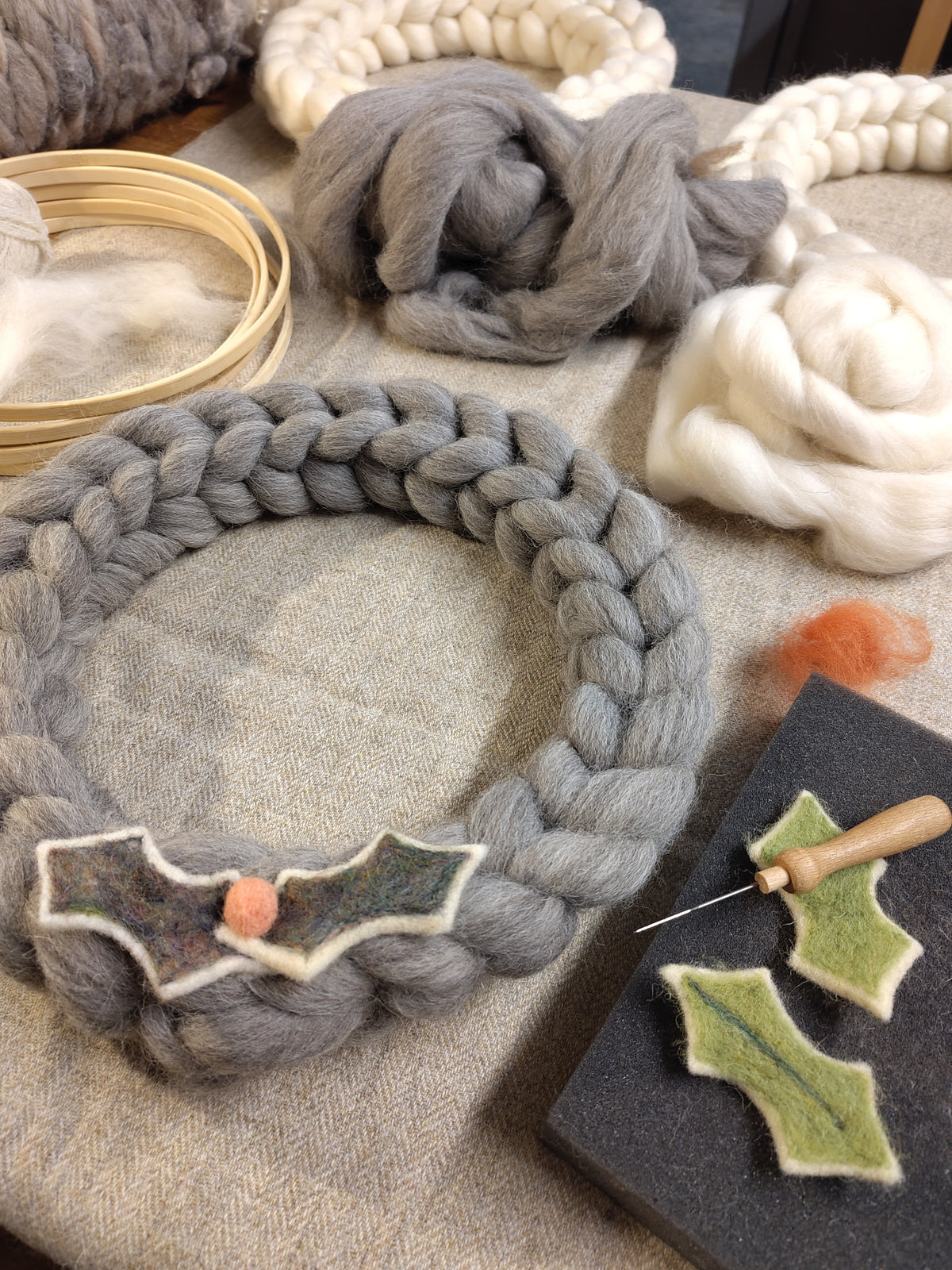 A braided gray wool wreath with a felted holly decoration, displayed on a textured fabric surface alongside wool roving, wooden rings, a needle, and green felted holly pieces.
